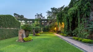 Un jardín exuberante con césped bien cuidado, setos recortados y altos muros de piedra cubiertos de hiedra: perfecto para eventos y conferencias. Al fondo se alza un antiguo edificio de piedra con ventanas arqueadas, suavemente iluminado por luces cálidas a lo largo de un sendero.