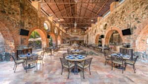 Un espacioso restaurante en uno de los resorts más exclusivos de México, con paredes de piedra, grandes arcos de ladrillo y ventanas redondas. Mesas y sillas de metal están ordenadas bajo un techo abovedado de madera, lo que lo hace perfecto para reuniones o eventos de conferencias.