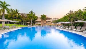 Una gran piscina al aire libre rodeada de tumbonas y sombrillas, con palmeras y vegetación de fondo, perfecta para relajarse en los hoteles de México o desconectar después de eventos de conferencias en los complejos turísticos cercanos bajo un cielo despejado.