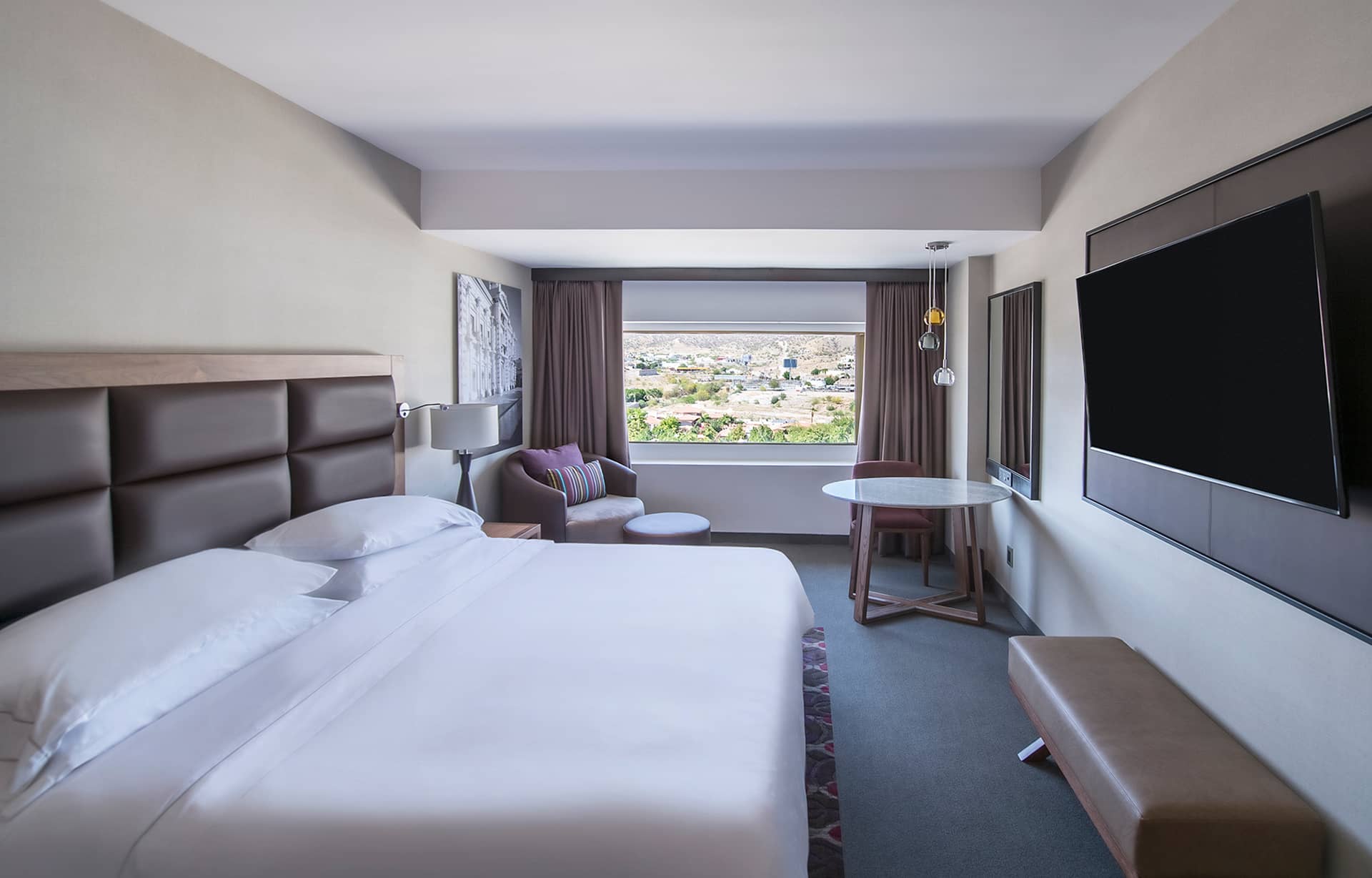A modern hotel room with a large bed, wall-mounted TV, small table and chairs by the window, armchair, and bench—perfect for relaxing after conference events or meetings. The window offers a cityscape view in the background.