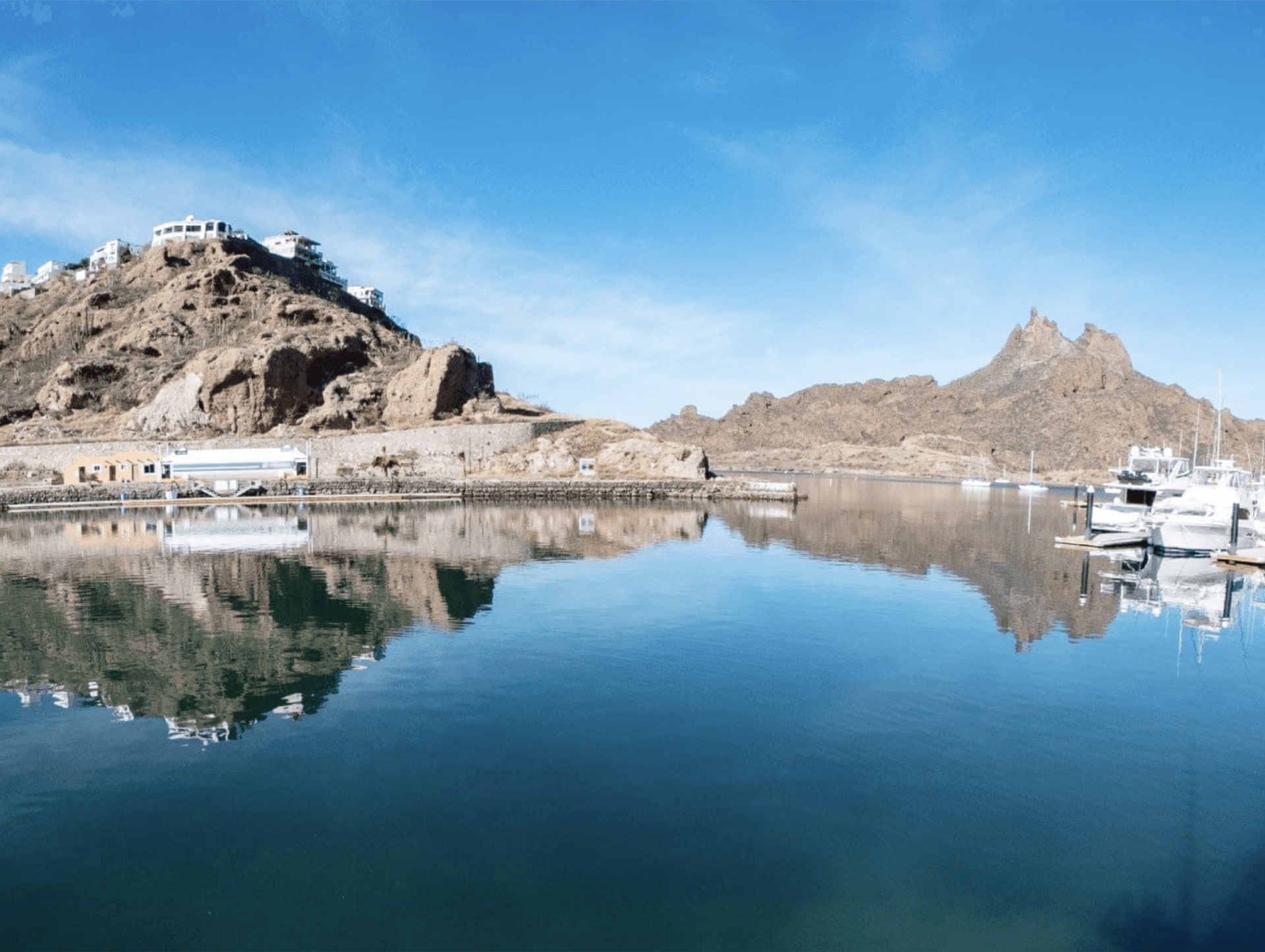 A calm marina with clear blue water reflecting rocky hills, boats, and white buildings under a bright blue sky—an inviting scene near top Mexico resorts and hotels, perfect for conference events or relaxing after meetings.