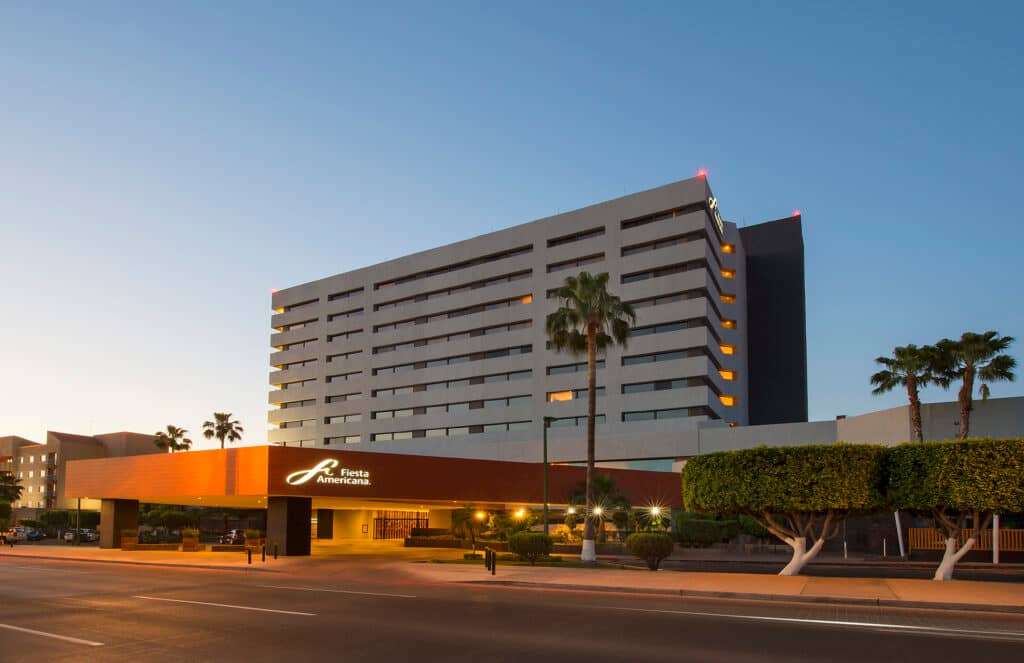 A modern multi-story hotel building with the sign Fiesta Americana is seen at sunset, perfect for meetings and conference events, with palm trees and neatly trimmed bushes in front, under a clear blue sky.