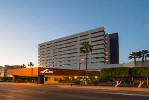 Un moderno hotel mexicano de varias plantas con techo plano y balcones, que cuenta con una imponente marquesina en la entrada y palmeras al frente, fotografiado al atardecer con el cielo tiñéndose de azul; ideal para reuniones o eventos de conferencias.