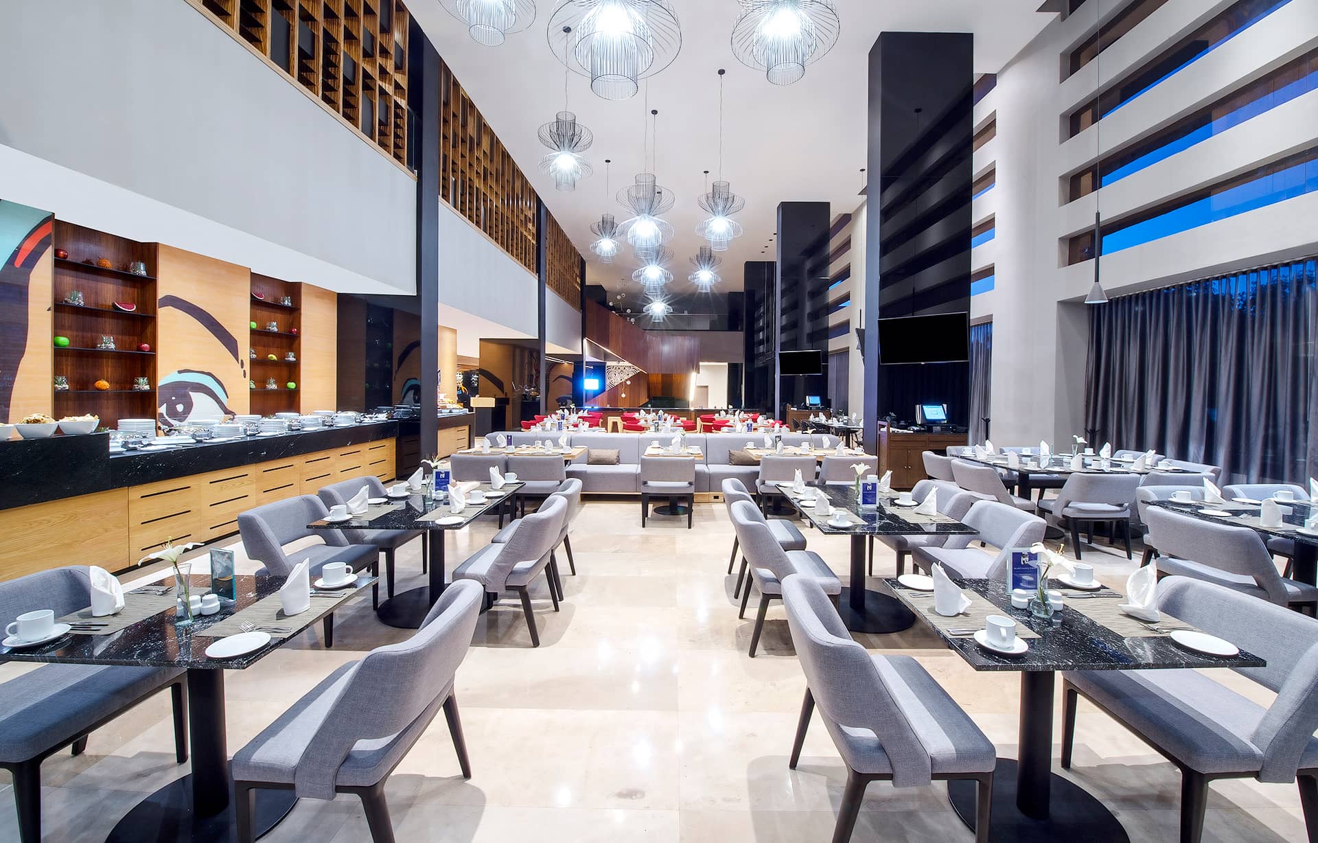 Modern, spacious restaurant perfect for conference events, with neatly arranged tables and grey chairs, elegant lighting fixtures, floor-to-ceiling windows with dark curtains, and a buffet area on the left with stylish decor.