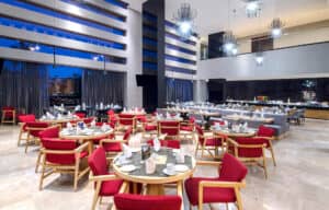 Un restaurante moderno y espacioso con mesas redondas, sillas rojas acolchadas, grandes ventanales, iluminación contemporánea y área de buffet. Ideal para reuniones o conferencias en hoteles y resorts de México. El ambiente es elegante y luminoso.