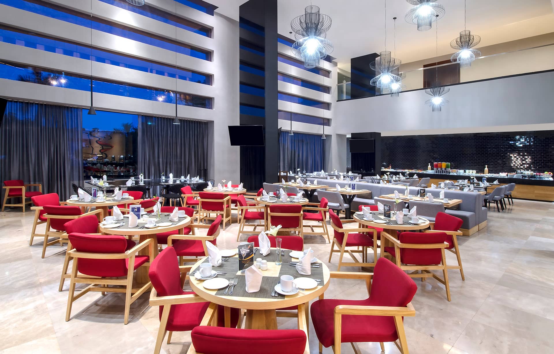 A modern, spacious restaurant with round tables set for dining, red cushioned chairs, large windows, contemporary light fixtures, and a buffet area. Ideal for meetings or conference events at Mexico hotels and resorts. The atmosphere is elegant and well-lit.