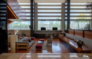 Un moderno vestíbulo de hotel con grandes ventanales, una zona de estar con sofás y sillones, una larga recepción de madera y un bar con botellas. La luz natural inunda el espacio, realzando la decoración contemporánea, ideal para reuniones o conferencias.