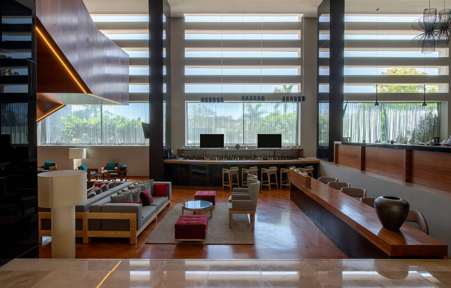 A modern hotel lobby with large windows, a seating area with sofas and chairs, a long wooden reception desk, and a bar lined with bottles. Natural light fills the space, highlighting contemporary decor ideal for meetings or conference events.
