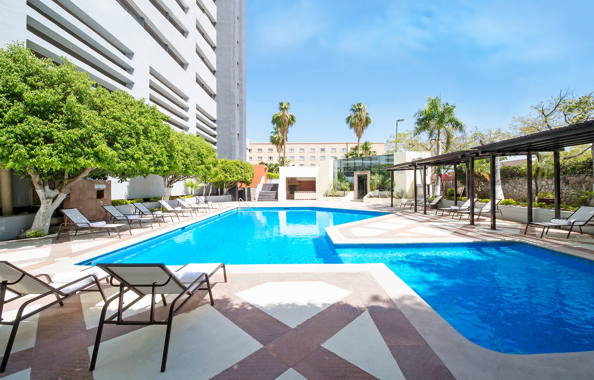 Outdoor swimming pool surrounded by lounge chairs, trees, and modern buildings at one of Mexico's top resorts, perfect for conference events or meetings, with clear blue sky overhead and palm trees in the background.