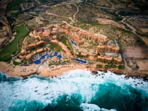 Vista aérea de un gran complejo turístico costero en México con múltiples edificios, piscinas y palmeras a lo largo de una playa de arena, perfecto para eventos y reuniones, bordeado por olas oceánicas de color turquesa y un terreno accidentado al fondo.