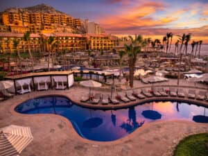 Un lujoso resort en México con piscina curva, tumbonas, cabañas y palmeras al atardecer. El vibrante cielo y las vistas al océano crean un escenario ideal para eventos de conferencias o reuniones de negocios inolvidables.