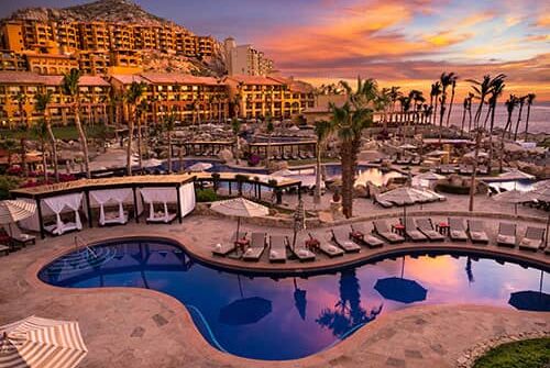 Luxury resort in Mexico with palm trees, pools, and lounge chairs at sunset, set against mountainous terrain and colorful sky, with large hotel buildings perfect for meetings and conference events in the background.