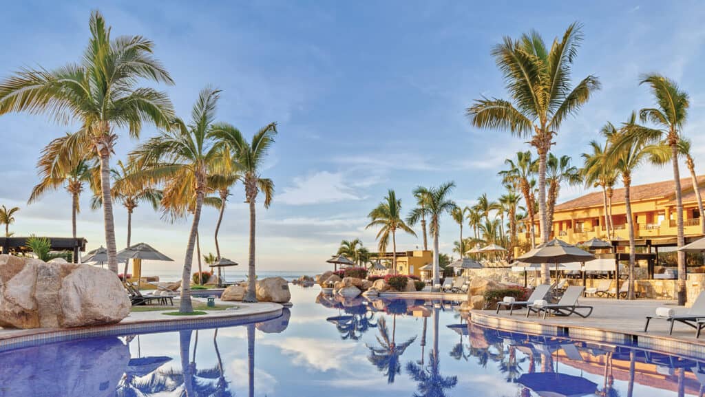A luxurious resort pool surrounded by palm trees, lounge chairs, and umbrellas, with clear blue sky and resort buildings in the background—an ideal setting for meetings or conference events at top Mexico hotels.