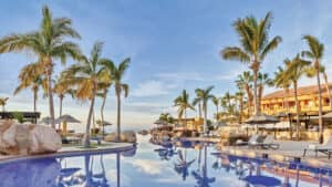 Una lujosa piscina de resort rodeada de palmeras, tumbonas y sombrillas, con un cielo azul despejado y los edificios del resort al fondo: un escenario ideal para reuniones o eventos de conferencias en los mejores hoteles de México.