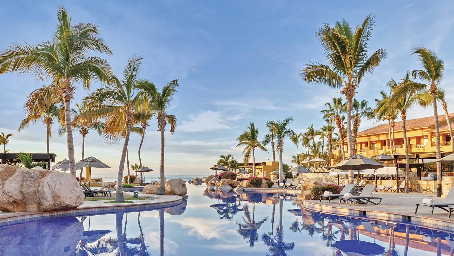 Una lujosa piscina de resort rodeada de palmeras, tumbonas y sombrillas, con un cielo azul despejado y los edificios del resort al fondo: un escenario ideal para reuniones o eventos de conferencias en los mejores hoteles de México.