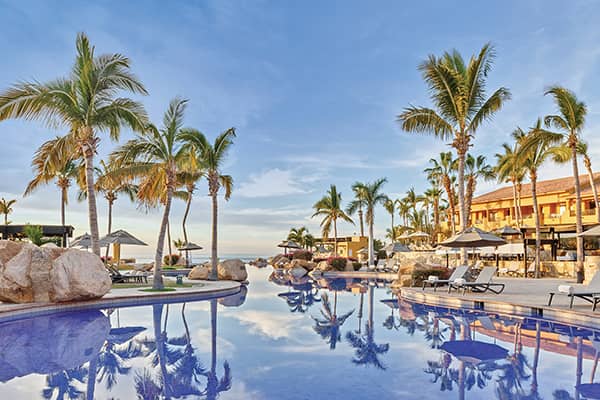 A tropical resort in Mexico with tall palm trees reflected in a clear swimming pool, surrounded by lounge chairs and umbrellas—perfect for meetings or conference events against a backdrop of a yellow hotel and blue sky.