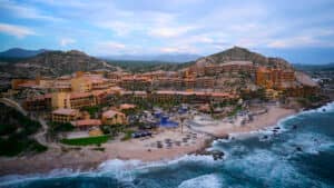 Vista aérea de un gran complejo turístico costero mexicano con edificios de color terracota, piscinas, palmeras y tumbonas en una playa de arena, ideal para reuniones o eventos de conferencias, con colinas rocosas de fondo y bordeado por las olas del océano.