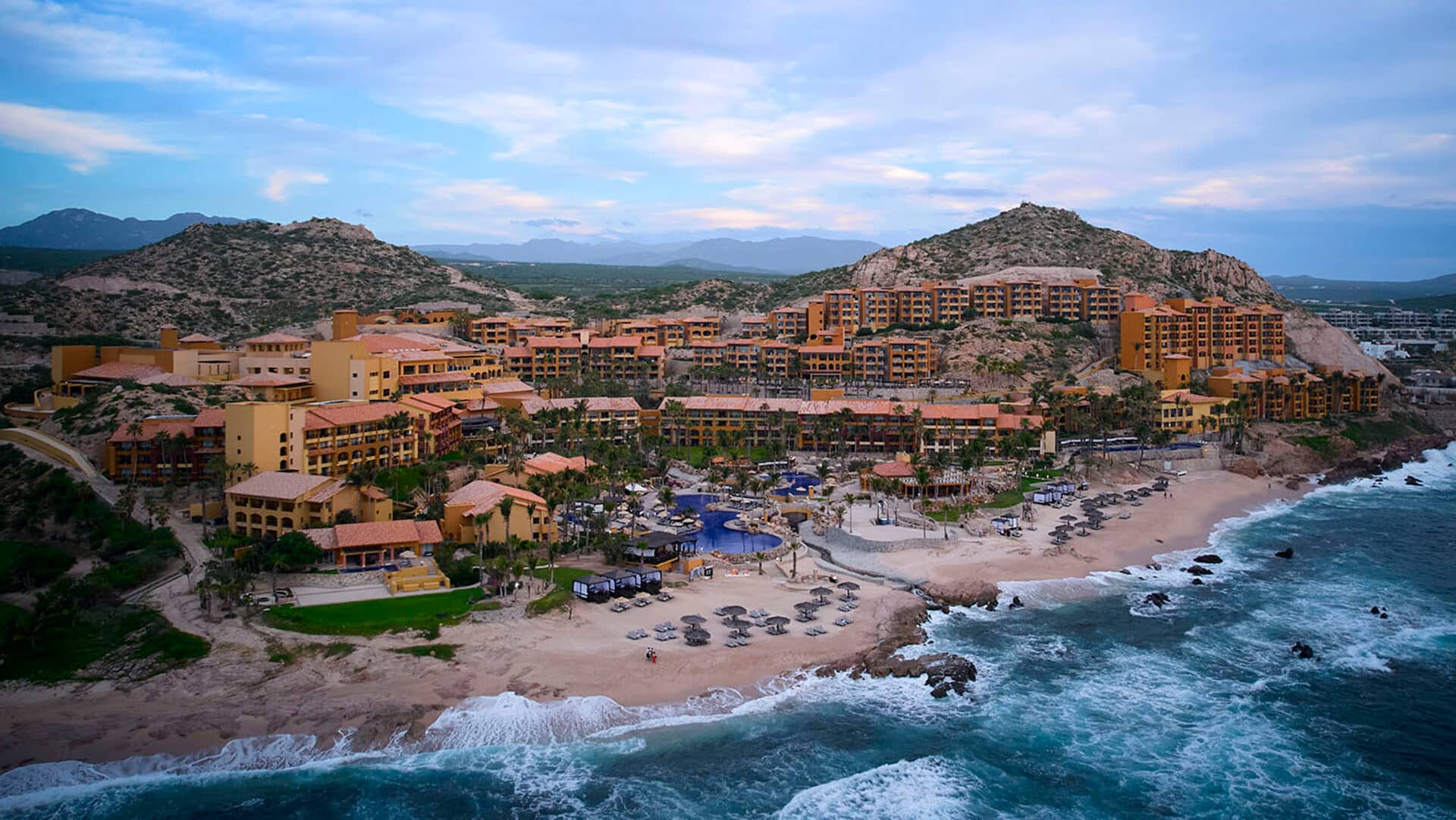 Vista aérea de un gran complejo turístico costero mexicano con edificios de color terracota, piscinas, palmeras y tumbonas en una playa de arena, ideal para reuniones o eventos de conferencias, con colinas rocosas de fondo y bordeado por las olas del océano.