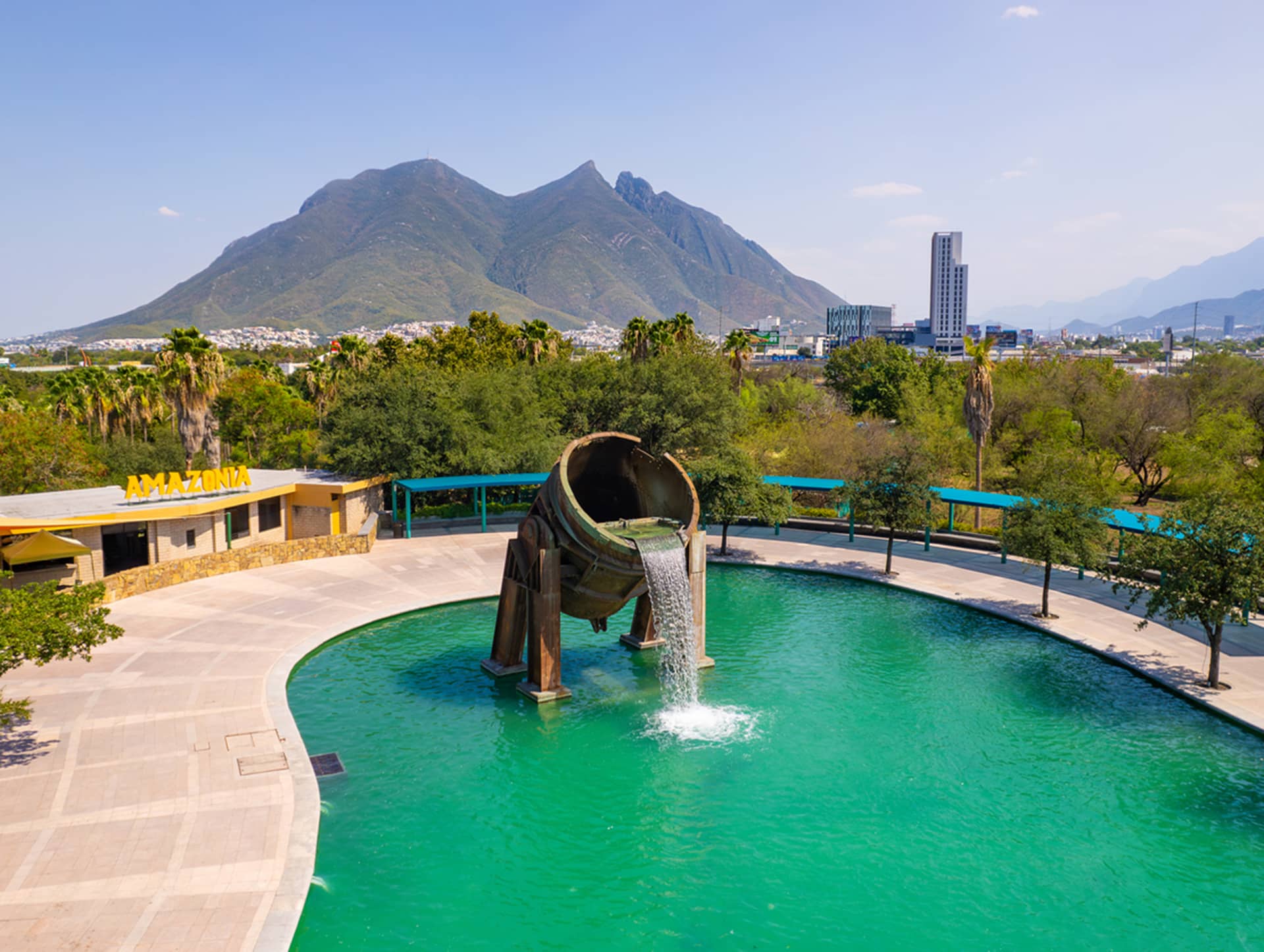 A large metal sculpture of a tilted foundry pot pours water into a circular pool, surrounded by trees. In the background, city buildings, resorts, and a mountain range are visible under a clear sky.