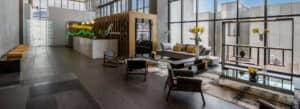 Un moderno vestíbulo de oficinas con techos altos, grandes ventanales, mobiliario contemporáneo, mostrador de recepción y flores amarillas decorativas sobre las mesas. La luz natural inunda el espacio, ideal para reuniones, y desde las ventanas se divisan los edificios de la ciudad.