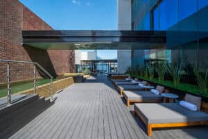 Moderna terraza al aire libre en uno de los resorts más exclusivos de México, ideal para reuniones o eventos corporativos. Cuenta con tumbonas, toallas blancas enrolladas, plantas en macetas, paredes de cristal y una barandilla metálica, junto a un elegante edificio de ladrillo y cristal, bajo un cielo despejado.