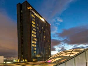 Un alto y moderno edificio hotelero con el letrero de Presidente InterContinental se ilumina al anochecer. Ideal para reuniones y conferencias, el hotel se alza junto a una estructura de cristal en forma de cúpula bajo un cielo azul y naranja.