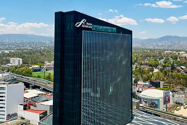 Tall glass skyscraper with Fiesta Americana sign on top, surrounded by city buildings, roads, and greenery—ideal for meetings or conference events—with mountains and a cloudy sky in the background.