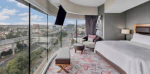 Una moderna habitación de hotel en uno de los mejores complejos turísticos de México, con una cama grande, sillón y otomana, y ventanales curvos que van del suelo al techo y ofrecen una vista panorámica de la ciudad: un refugio ideal después de reuniones o conferencias exitosas.