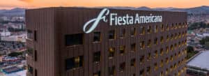 Un hotel Fiesta Americana de varias plantas, de color marrón, al atardecer, con el paisaje urbano y las montañas de fondo. El gran logotipo y el nombre del hotel, en blanco, destacan prominentemente, lo que lo convierte en una opción ideal para reuniones o conferencias en México.