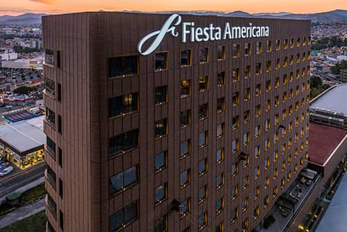 Tall brown hotel building with many windows, illuminated at dusk. The Fiesta Americana sign is displayed at the top. Ideal for conferences and meetings, this Mexico hotel offers stunning cityscape and mountain views in the background.