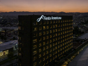 Un alto edificio iluminado del hotel Fiesta Americana al anochecer, con luces cálidas que brillan desde sus ventanas —un punto de referencia acogedor para eventos de conferencias—, con el paisaje urbano y la silueta de las montañas de fondo bajo un colorido cielo de puesta de sol.