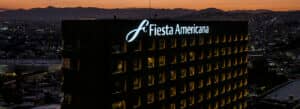 Un imponente edificio del hotel Fiesta Americana se ilumina al anochecer, creando un ambiente perfecto para conferencias y reuniones, con las luces de la ciudad y las montañas visibles al fondo bajo un cielo anaranjado.