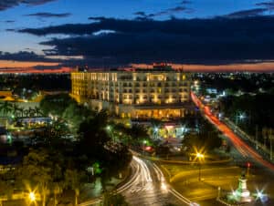 Un gran hotel iluminado se alza en una concurrida intersección al anochecer, ideal para eventos y conferencias. Las luces de los coches serpentean por las calles, con árboles y un monumento en primer plano y un colorido cielo de atardecer al fondo.