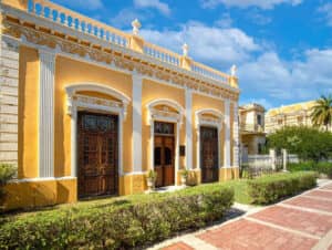 Un edificio histórico de estilo colonial, de color amarillo y blanco, con detalles ornamentados, grandes ventanales arqueados con rejas de hierro forjado y plantas en macetas en la entrada: un entorno ideal para celebrar conferencias o reuniones memorables en México.