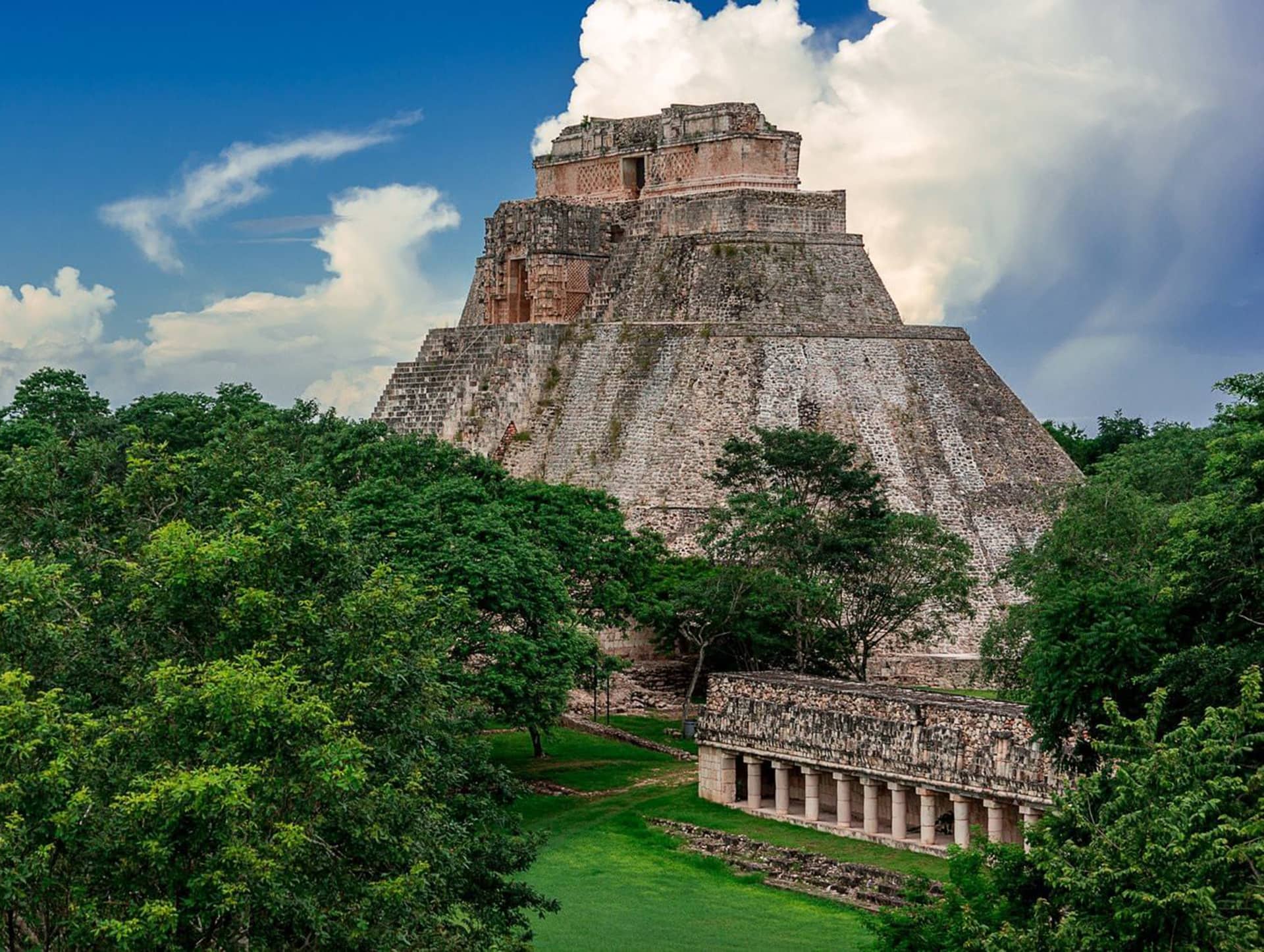 Ancient Mayan pyramid rises above lush green trees and grass, with a blue sky and clouds in the background; a stone building with columns sits beside the pyramid, evoking the timeless elegance of Mexico hotels and resorts.