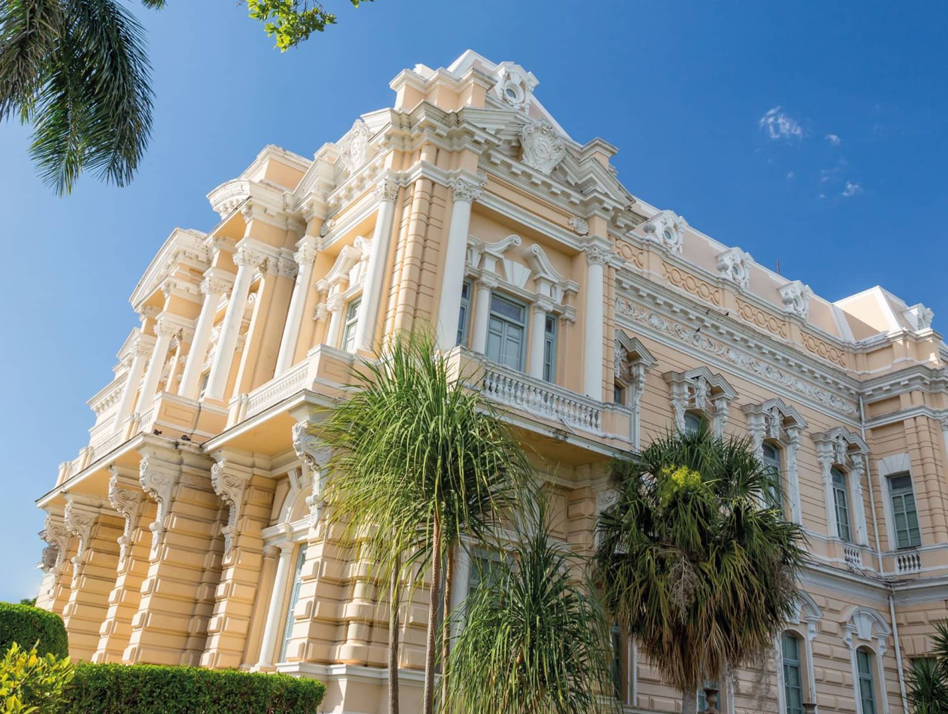 A grand, ornate beige mansion with tall columns, decorative moldings, and large windows—perfect for conference events or meetings—surrounded by palm trees under a clear blue sky.
