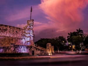 Un gran monumento de piedra con intrincadas tallas se alza en una rotonda bajo un espectacular y colorido atardecer. Árboles y luces de coches borrosas rodean el monumento, cerca de populares hoteles y centros de convenciones en México, con un letrero de McDonald's al fondo.