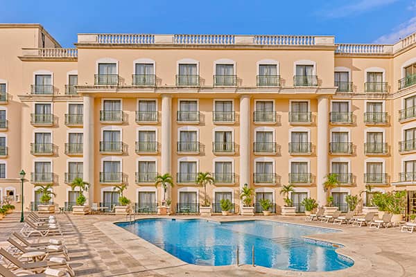 A four-story beige hotel with balconies overlooks a kidney-shaped outdoor swimming pool, perfect for relaxing after conference events or meetings at one of Mexico’s premier resorts, surrounded by lounge chairs and small palm trees under a clear blue sky.