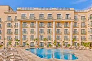 Hotel de cuatro plantas de color beige con balcones que dan a una piscina, perfecto para reuniones y eventos, rodeado de tumbonas y palmeras bajo un cielo azul despejado.