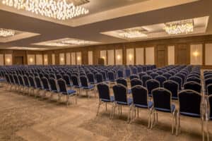 En este complejo turístico mexicano, una amplia sala de conferencias vacía está acondicionada con filas de sillas azules frente a un escenario. Elegantes lámparas de araña y cálidas luces de pared realzan el ambiente espacioso y formal, ideal para reuniones o eventos.