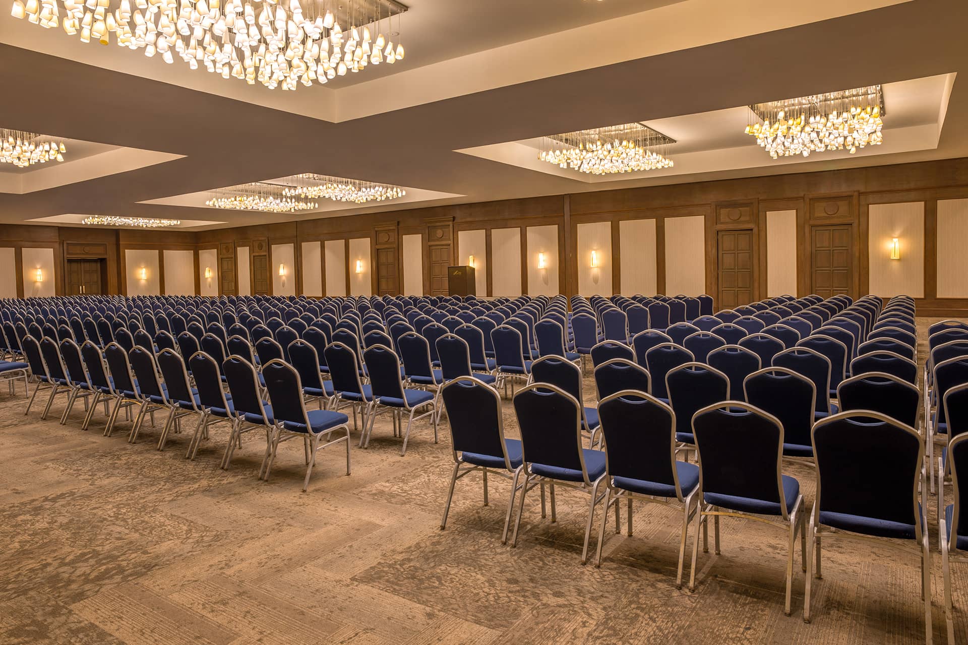 A large, empty conference room at this Mexico resort is set up with rows of blue chairs facing a stage. Elegant chandeliers and warm wall lights enhance the spacious, formal atmosphere—ideal for meetings or conference events.