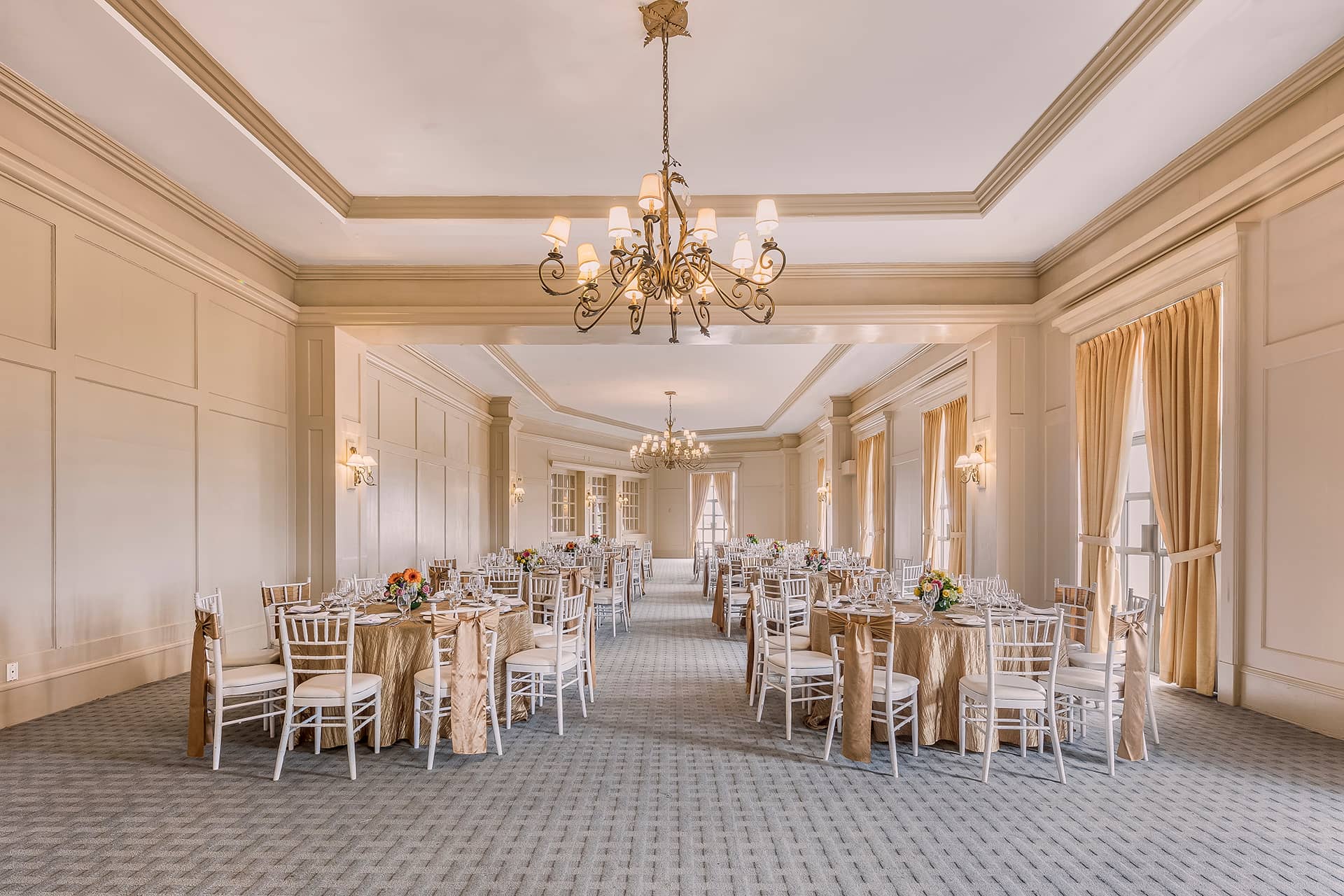 A spacious, elegant banquet hall ideal for conference events at resorts in Mexico, featuring chandeliers, gold tablecloths, white chairs, floral centerpieces, and large windows with gold curtains that fill the room with natural light.