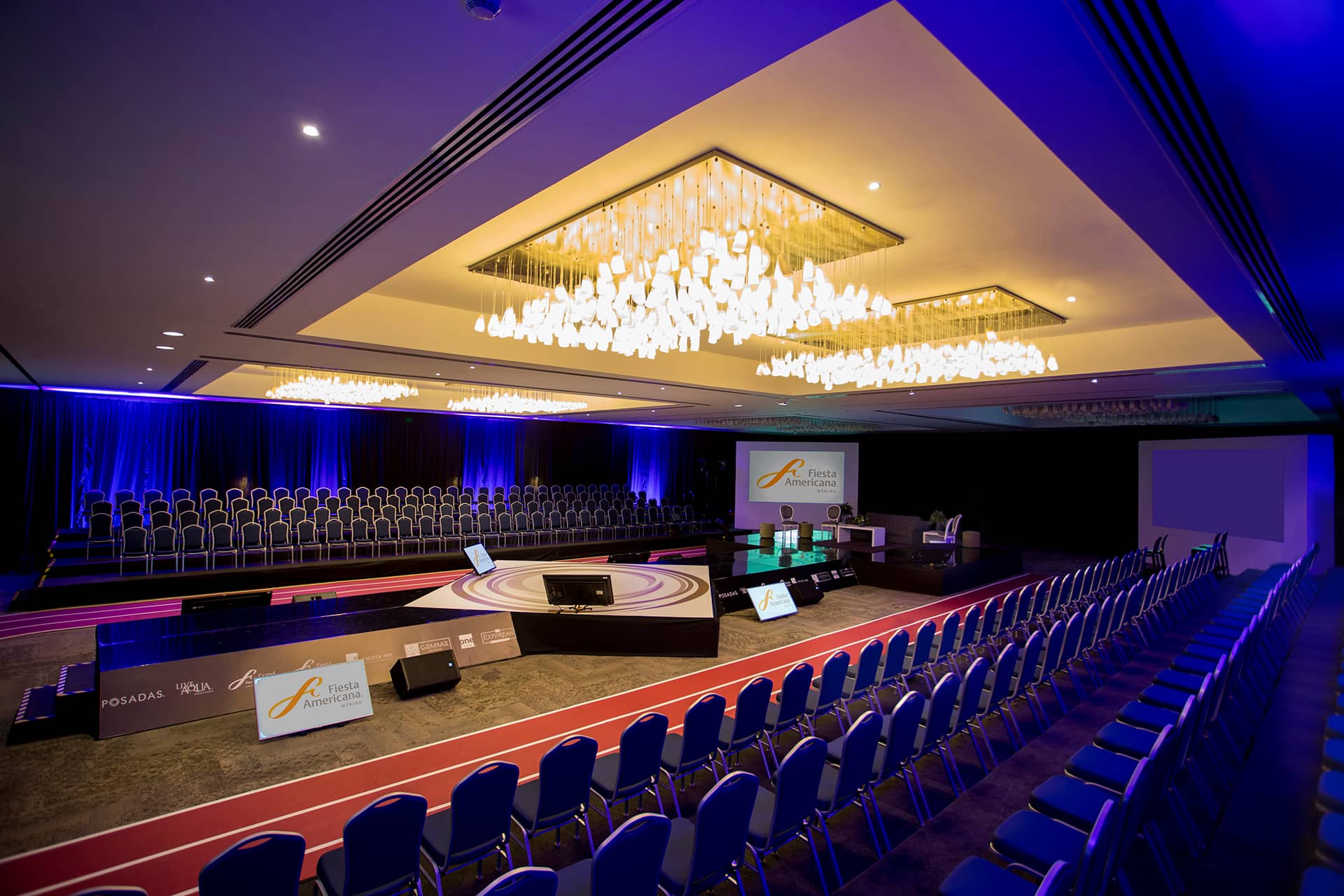 Large, modern conference room at one of Mexico's premier resorts, with rows of blue chairs around a central stage. Elegant chandeliers, purple accent lighting, and presentation screens create an ideal setting for meetings or conference events.