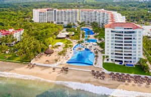 Vista aérea de un complejo turístico frente al mar en México con dos grandes edificios blancos, piscinas, palmeras y sombrillas de paja que bordean la orilla; ideal para eventos y reuniones en medio de una exuberante vegetación y las olas del océano.