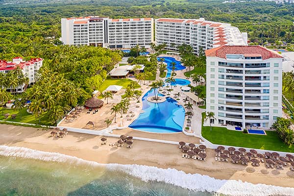 Aerial view of a beachfront resort in Mexico with two large white buildings, outdoor swimming pools, palm trees, and umbrellas on a sandy beach—perfect for meetings and conference events beside the ocean.