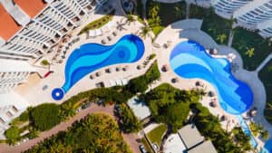 Vista aérea de dos piscinas azules de formas sinuosas en un moderno hotel resort de México, rodeadas de tumbonas, palmeras, jardines paisajísticos y aparcamiento cercano, ideal para reuniones o eventos de conferencias.