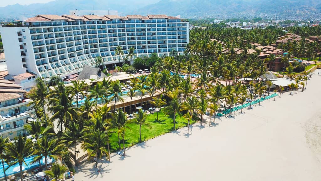 Aerial view of a beachfront resort in Mexico with a large, multi-story hotel, rows of palm trees, green lawns, and lounge chairs on a sandy beach—ideal for meetings and conference events, with mountains and a city in the background.