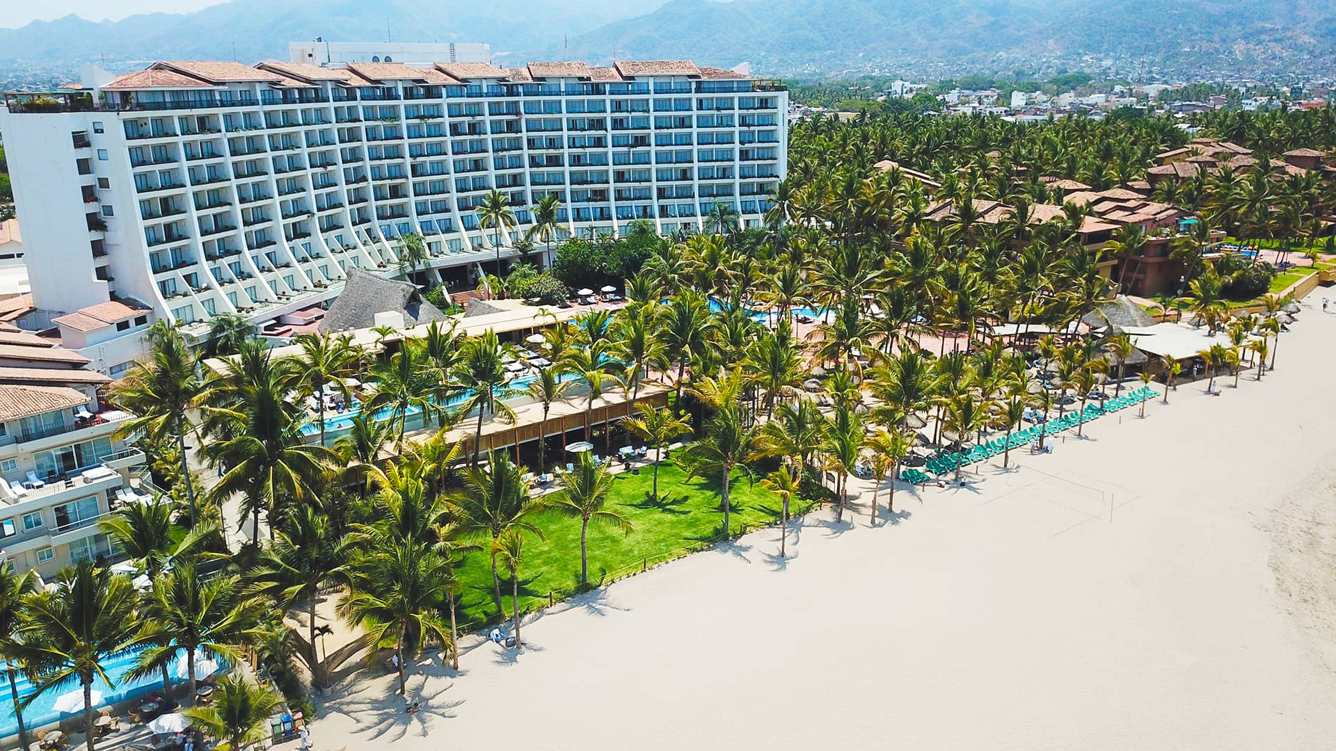 Vista aérea de un complejo turístico frente al mar en México, con un gran hotel de varias plantas, hileras de palmeras, césped verde y tumbonas en una playa de arena; ideal para reuniones y eventos, con montañas y una ciudad al fondo.