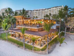 Un restaurante frente al mar con dos niveles al aire libre, rodeado de palmeras y con vistas a una playa de arena y a los imponentes hoteles turísticos mexicanos al atardecer. Los comensales disfrutan de la comida en mesas ubicadas en ambos niveles, ideales para reuniones o eventos.