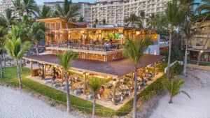 Un restaurante frente al mar con dos niveles de terraza, rodeado de palmeras y un ambiente animado. Al fondo, se alza un gran hotel de varios pisos en México, ideal para conferencias y reuniones memorables en un entorno tropical vibrante.